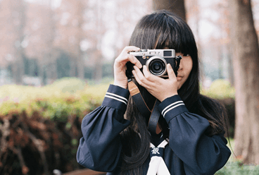 La imagen muestra a una chica japonesa de frente haciendo una foto con su cámara y detrás de ella hay árboles y mucha vegetación.