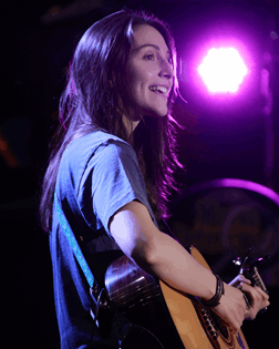 La imagen muestra a una chica con pelo largo que está de lado tocando una guitarra en un escenario.