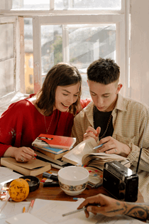 La imagen muestra dos jóvenes sentados ante varios libros, apuntes en actitud de trabajo y estudio.