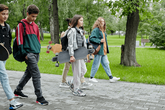 La imagen muestra un grupo de chicos y chicas jóvenes caminando por una senda en el parque, llevan mochilas a las espaldas y una joven porta un monopatín.