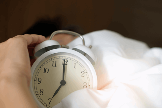 La imagen muestra las manos de alguien saliendo de unas sábanas blancas, están apagando la alarma de un despertador. 