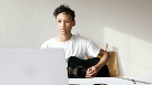 La imagen muestra un joven moreno con una guitarra en los brazos, sentado frente a su ordenador, que está mirando. 