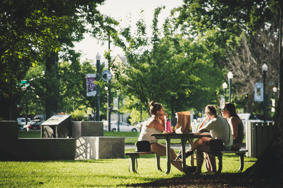 La imagen muestra a tres chicas jóvenes sentadas a una mesa en un gran parque, están comiendo. 