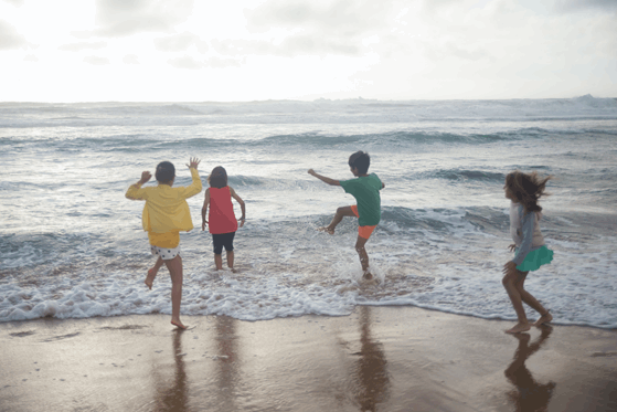  La imagen muestra un grupo de niños y niñas por detrás, saltando olas a la orilla del mar al atardecer. 