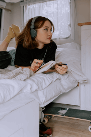 La imagen muestra a una chica joven tumbada boca abajo en su cama, lleva unos cascos para escuchar música y está escribiendo en un cuaderno, debajo de la cama se ve un monopatín.