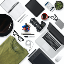 La imagen muestra un conjunto variado de objetos colocados sobre una superficie: una camiseta verde, una tablet, un sombrero, unas gafas, unos auriculares, un ordenador portátil, una libreta, un lapicero, una cámara, una taza de café, un móvil y una pequeña maceta.
