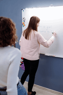  La imagen muestra a una chica escribiendo y explicando en una pizarra al resto de sus compañeros de clase.