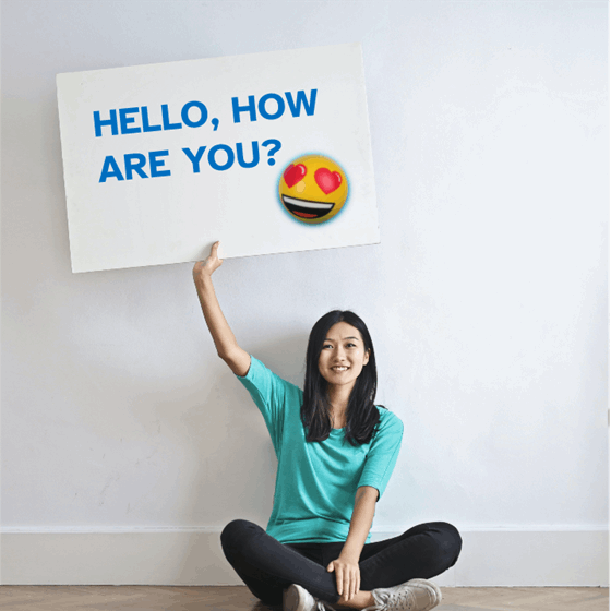  La imagen muestra una chica sentada sosteniendo un gran cartel con su brazo derecho en el que se ve la frase “hola, cómo estás”, junto a un emoji sonriente con los ojos de corazones.