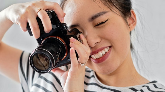 La imagen muestra a una chica con una cámara en las manos, y ajustada al ojo, en posición de disparar una foto.