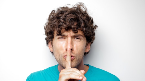 La imagen muestra un joven con el dedo índice de su mano derecha levantado tapándose los labios con él, indicando silencio.