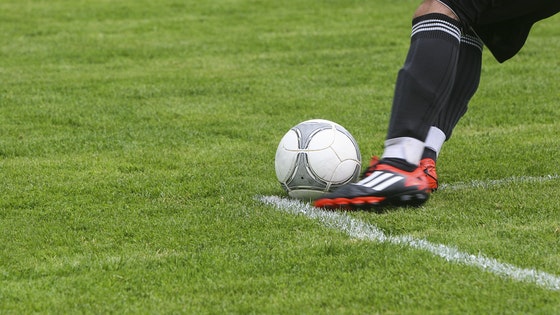 La imagen muestra los pies de un futbolista con el balón en el terreno de juego. 