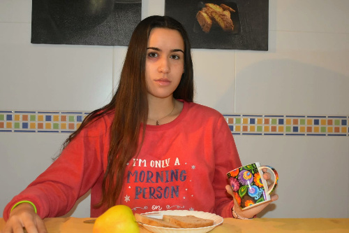 La foto muestra una chica con el ojo irritado en una cocina y delante de su desayuno.