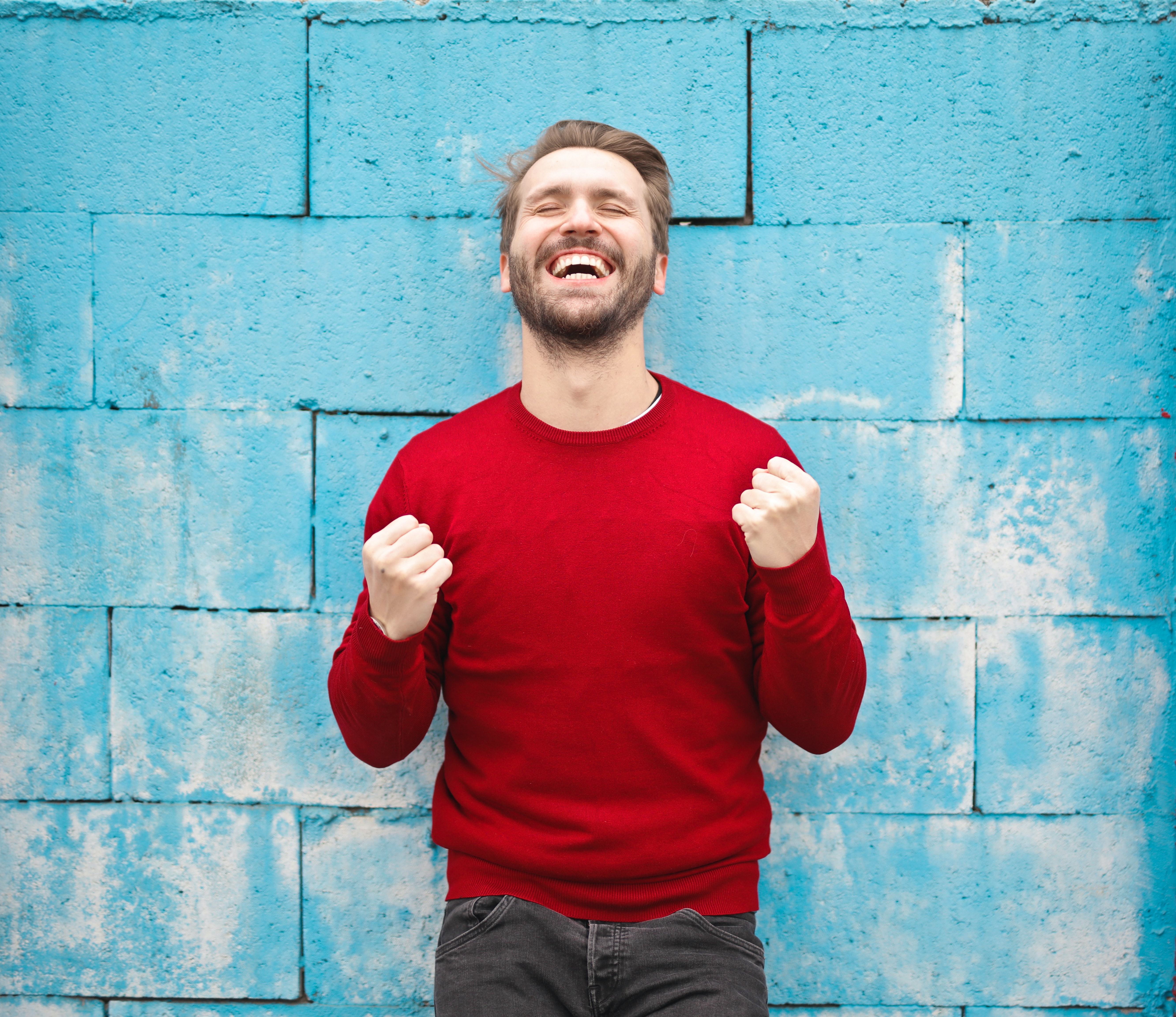 En la imagen se muestra un hombre con un jersey rojo y los puños cerrados delante de un muro de ladrillos azules mostrando satisfacción con una expresión de alegría