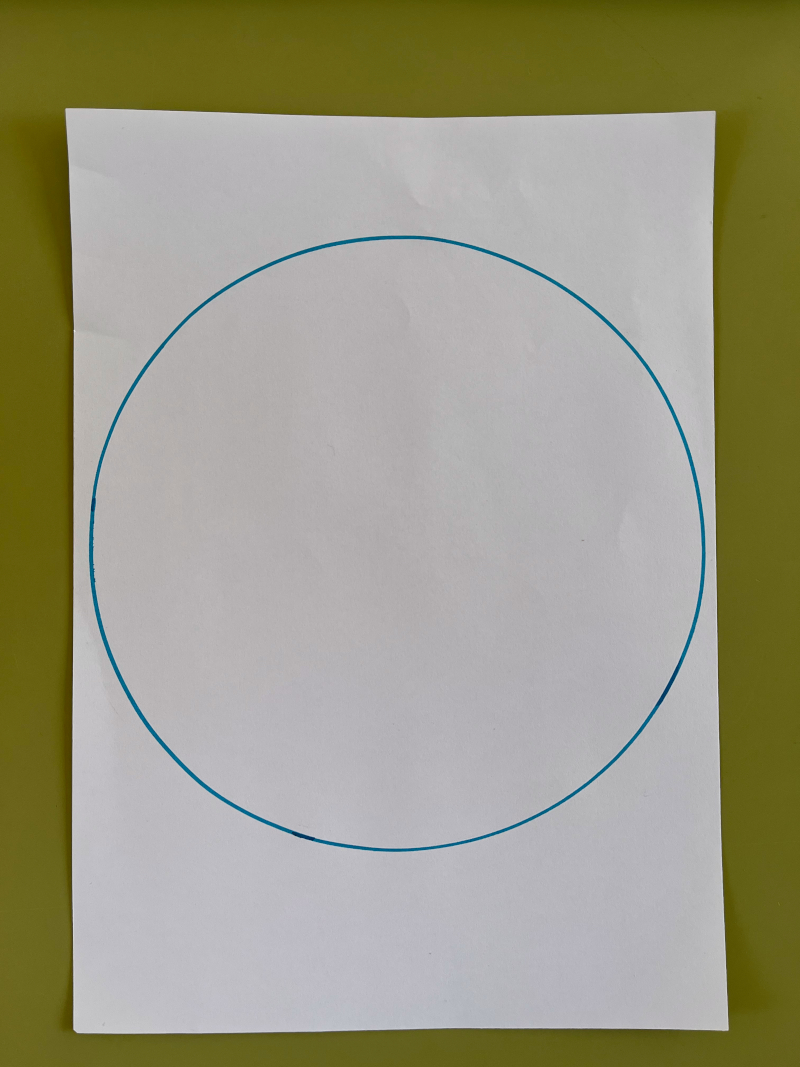 En la imagen puedes ver un círculo azul pintado en un folio en blanco, sobre una mesa escolar verde