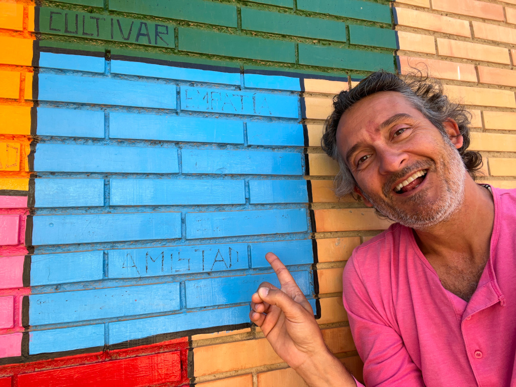 En la imagen puedes ver una persona sonriente señala un cuadro azul, de entre otras formas de distintos colores pintadas sobre una pared de ladrillo visto