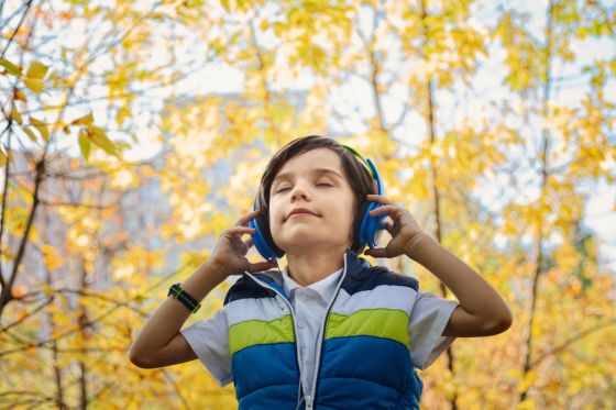 Se puede ver un niño con unos cascos escuchando música en un parque. Detrás del niño se aprecia árboles con las hojas anaranjadas y marrones en señal de estar en la estación de otoño.