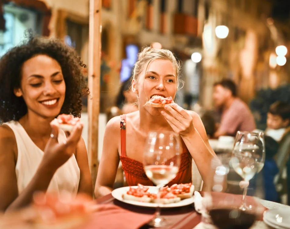 La imagen muestra a dos personas cenando en una mesa de una terraza. Una de ellas se está llevando comida a la boca y la otra mira la comida que tiene en su mano. Las dos sonríen.