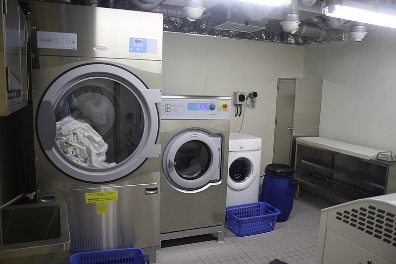 La imagen muestra tres lavadoras y dos cestos de la ropa azules. Una de ellas tiene ropa sucia dentro donde dos de ellas son lavadoras industriales de color plateado y una blanca a continuación. 