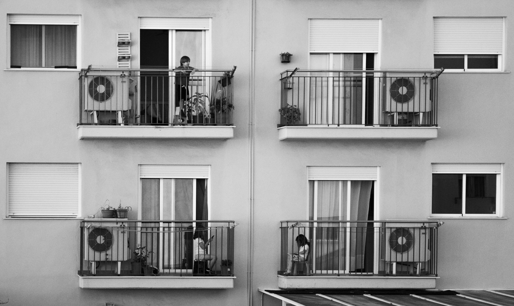 La imagen en blanco y negro muestra cuatro balcones de casas vecinas muy cercanos, y en tres de ellos hay un niño o niña.