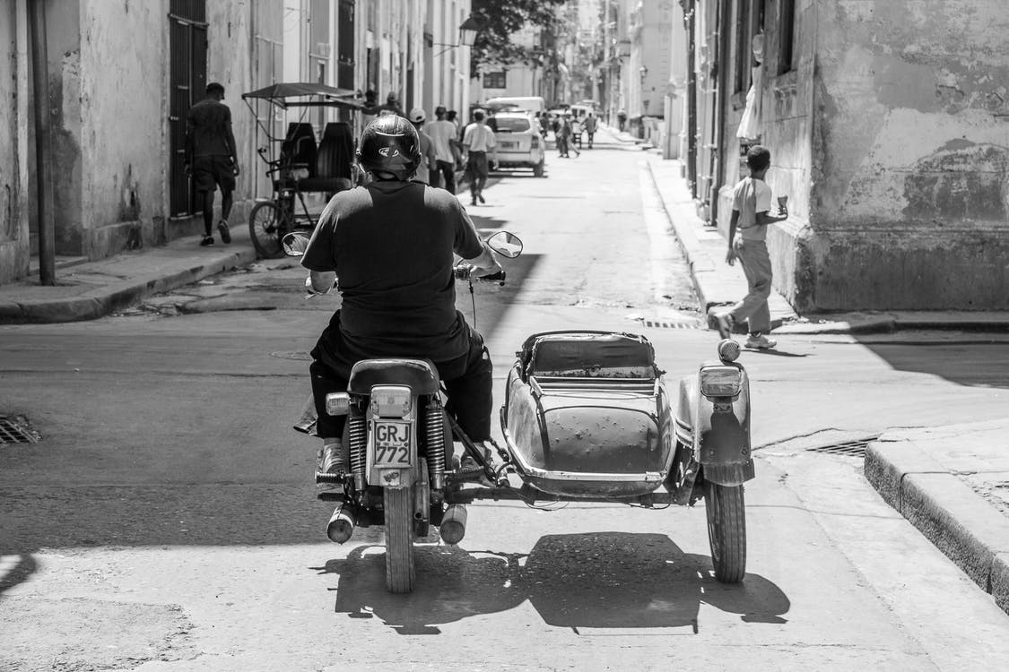 La imagen en blanco y negro muestra a un hombre de espaldas montado sobre una moto antigua circulando por una calle de una ciudad.