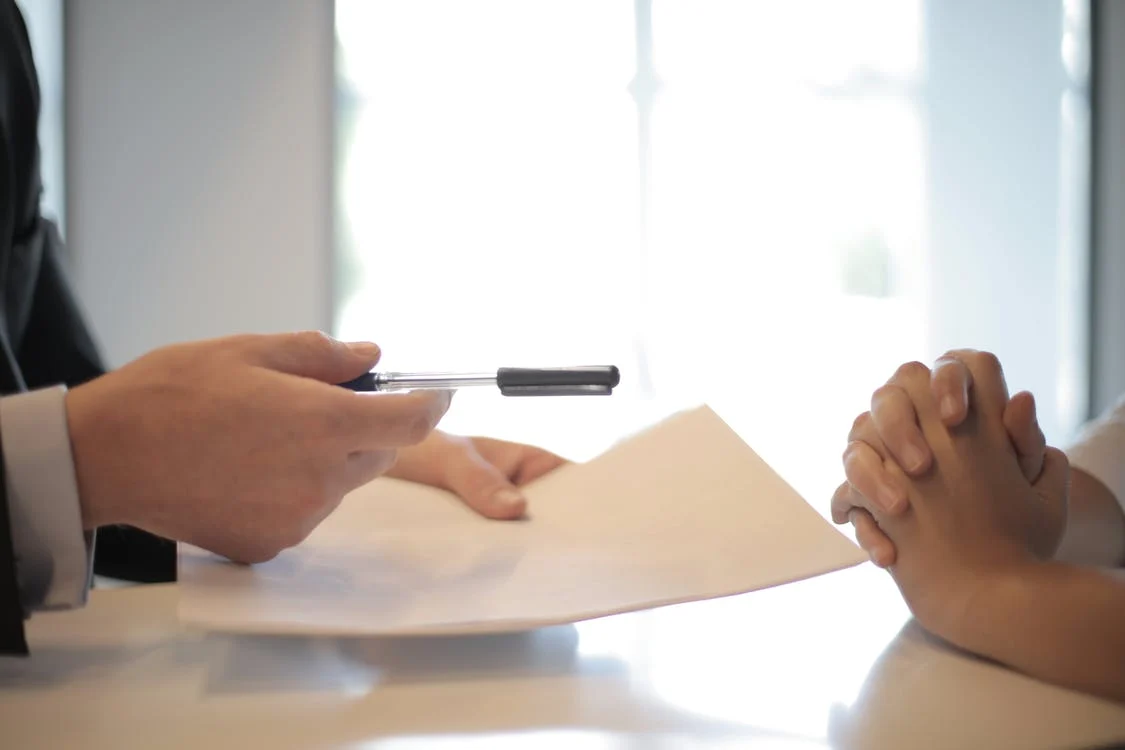 La imagen muestra una persona que sujeta en una mano un papel y en la otra un bolígrafo que da a otra persona para que lo firme.