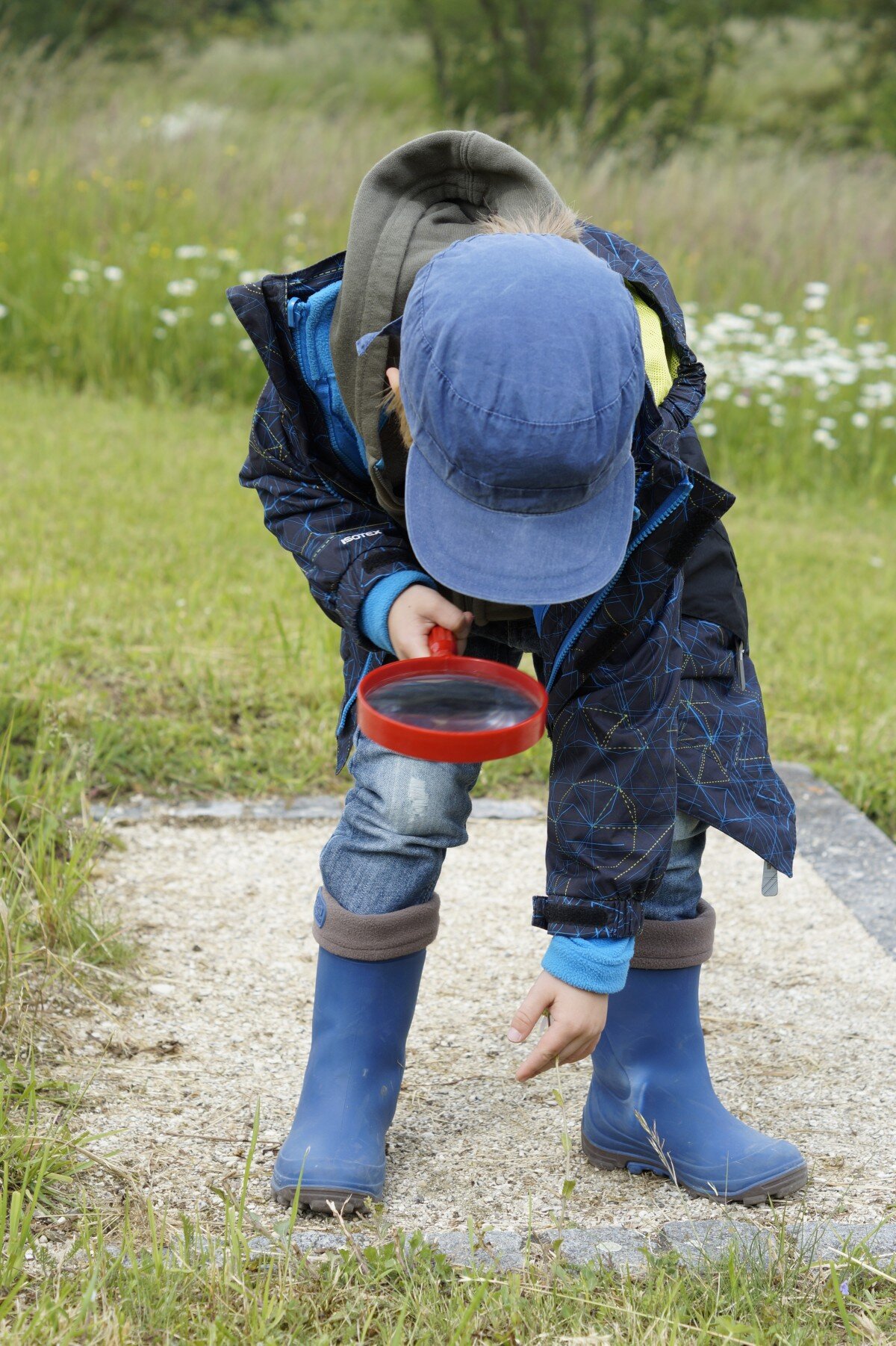 La imagen muestra un niño con una gorra azul agachado mientras observa algo en el suelo con una lupa. 