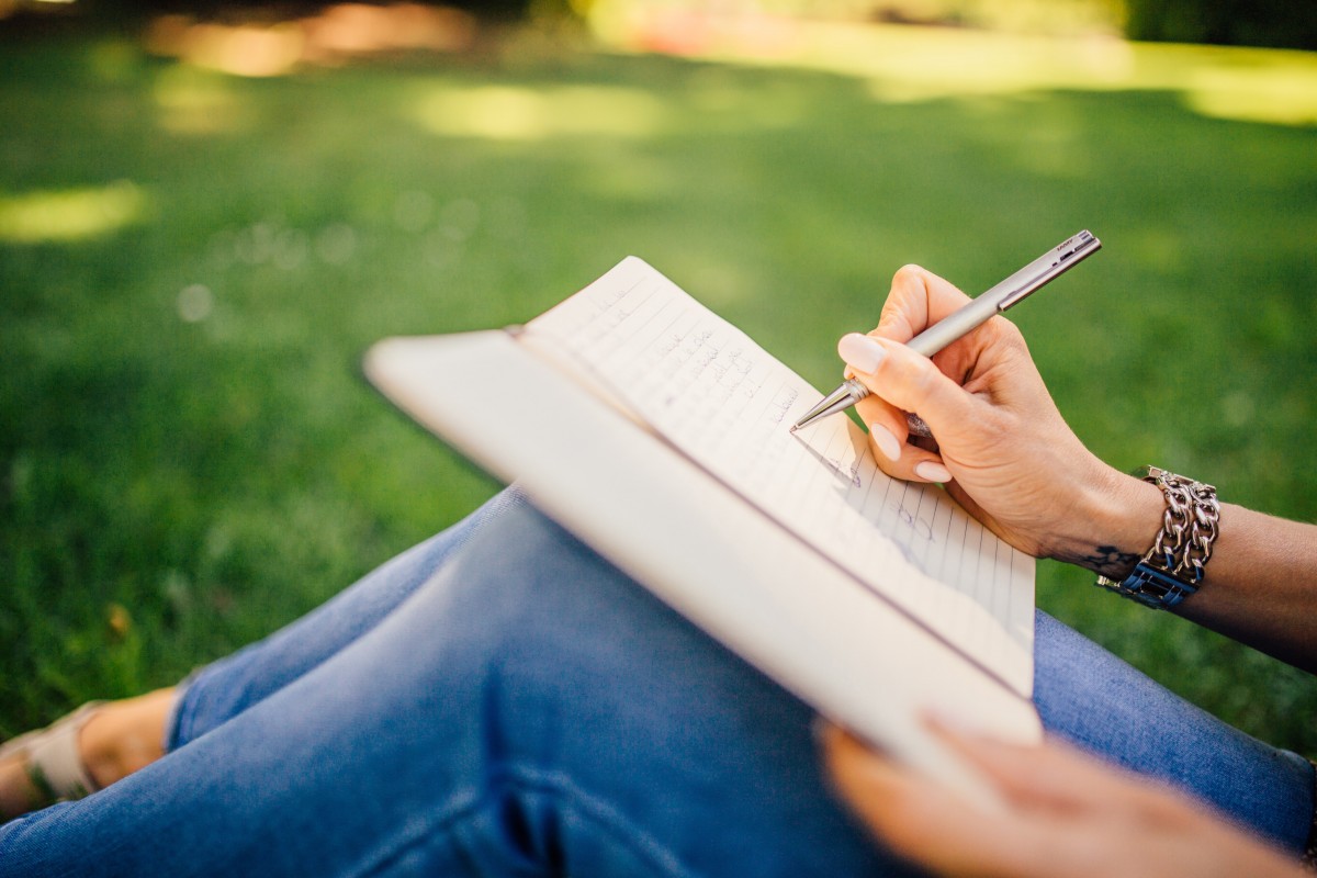 La imagen muestra a una persona sentada sobre la hierba mientras escribe en un cuaderno que tiene apoyado sobre las rodillas. 