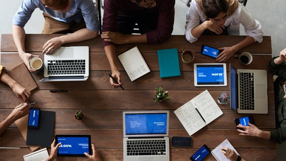 La imagen muestra un grupo de personas con ordenadores alrededor de una mesa. 