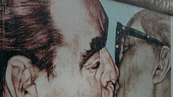  La imagen muestra un muro pintado con la imagen de dos hombres besándose en la boca y una persona pasando por delante.