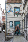 La imagen muestra una fachada de un edificio pintado con una escena de comic donde se ve un hombre colgado del tejado, una mujer intentando ayudarlo y debajo otro señor con un perro asustado por si le cae encima.