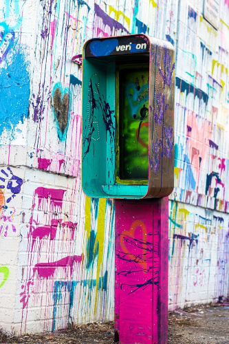  La imagen muestra una cabina de teléfonos con pintadas de muchos colores y detrás una pared con brochazos de pintura también de diversos colores.