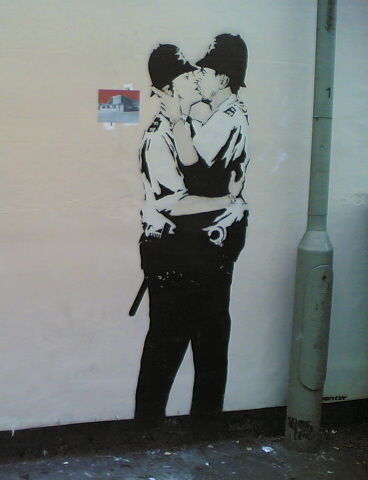La imagen muestra la obra de Banksy, en la que dos hombres policías se besan.