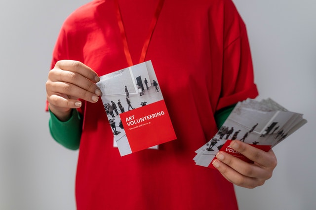 La imagen muestra a una persona con camiseta roja sosteniendo en su mano izquierda un puñado de folletos y, en su mano derecha, mostrando uno de ellos.