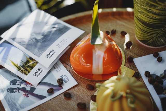 La imagen muestra tres folletos sobre una mesa de madera al lado de unos granos de café y una gran vela de color naranja.