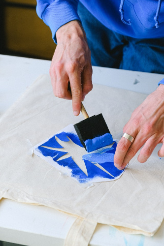 La imagen muestra a un artista utilizando una plantilla, brocha y pintura azul para crear una obra de arte.
