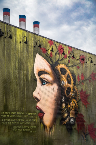 La imagen muestra un mural con una imagen de grandes dimensiones pintada en una pared. La imagen es la cara de una chica mitad humana mitad robot y con un mensaje escrito debajo.