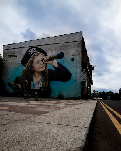 La imagen muestra un mural pintado en el lado de una pared de un edificio, en el que aparece una niña con sombrero pirata agarrando un osos de peluche y un catalejo.