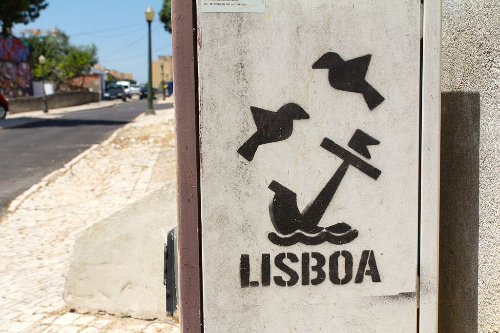 La imagen muestra una pintada en blanco y negro utilizando plantillas representando la ciudad de Lisboa con un ancla y dos pájaros.