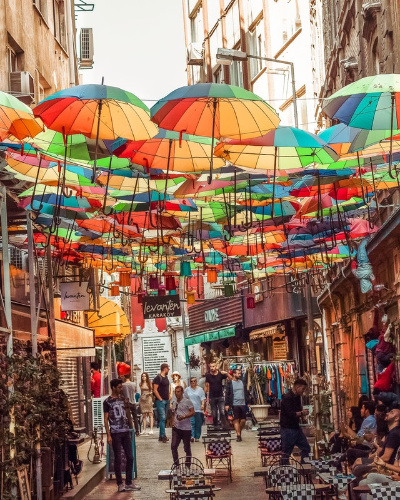 La imagen muestra una calle con multitud de paraguas de colores suspendidos en el aires, colgados de pared a pared.