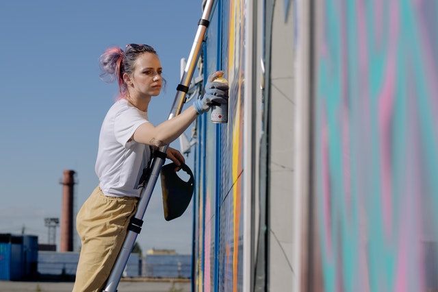 La imagen muestra a una artista urbana subida a una escalera pintando un graffiti con aerosol.
