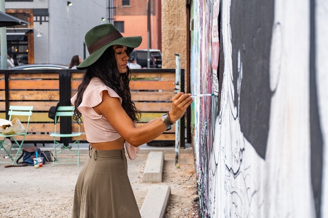 La imagen muestra a una artista urbana pintando con pincel un mural.