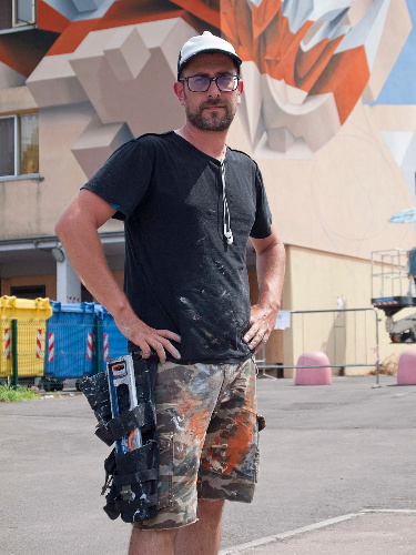 La imagen muestra a un artista urbano con manos y ropas manchadas de pintura y algunas herramientas en un cinturón de trabajo.