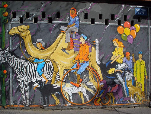 La imagen muestra un mural en donde aparecen caminando por una calle y semáforo en la esquina:  un hombre con turbante encima de un camello, una mujer con sombrero cowboy a lomos de una cebra, otra chica a lomos de un avestruz,un hombre montando una bicicleta de circo, un hombre con mono, pasamontañas y gafas con una rueda en las manos y ruedas en las rodillas, cuatro perros, un hombre con globos y otro con utensilios para barrer.