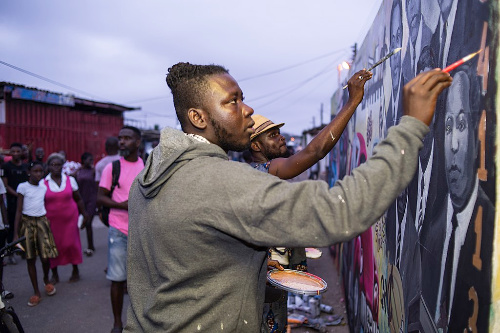 La imagen muestra un concurso de graffiti y unos artistas pintando en las paredes mientras hay gente mirando las pinturas.