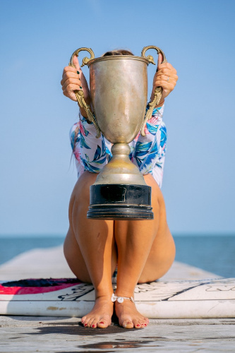 La imagen muestra a una joven sentada sobre una tabla de surf, levantando un trofeo. 