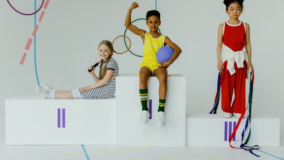 La imagen muestra tres niños en un podio, uno lleva una pelota, otro una raqueta y otro unas cintas de gimnasia rítmica. 