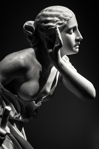 La imagen muestra la escultura de mármol blanco de una mujer mostrando un pecho al aire y con su mano izquierda al lado de su oreja izquierda a modo de escucha.