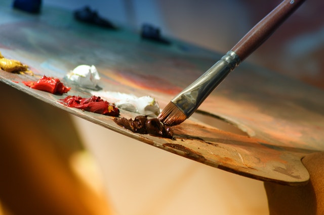 La imagen muestra un pincel cogiendo pintura de color marrón de una paleta con más pinturas.