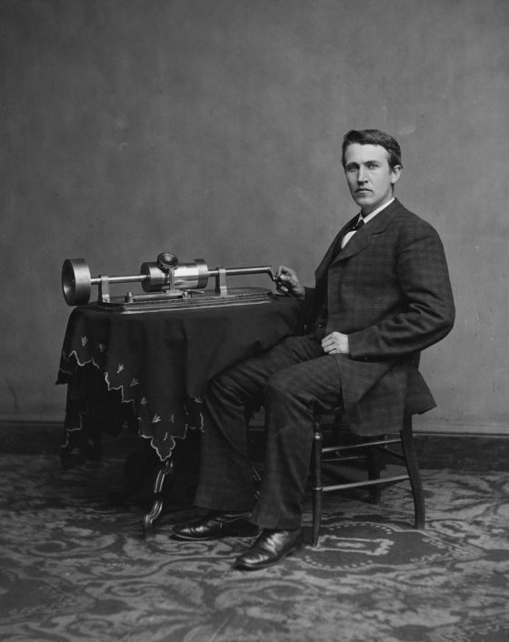 Es una imagen en blanco y negro en la que aparece un hombre sentado y junto a él una mesa en la que reposa un fonógrafo, invento que servía para grabar o reproducir sonidos