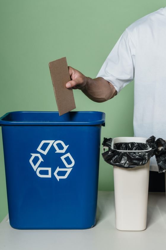 Aparece la mano de un hombre depositando un trozo de cartón en un contenedor azul con el símbolo del reciclaje, tres flechas blancas que forman un triángulo y que representan la idea de proceso continuo